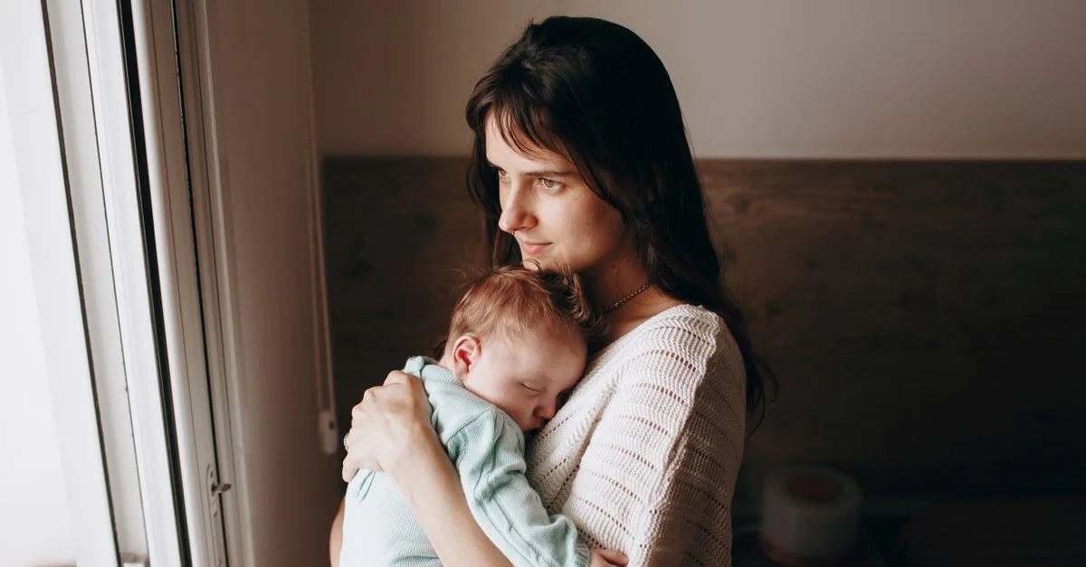 Mãe pensativa olhando pela janela enquanto o bebê dorme